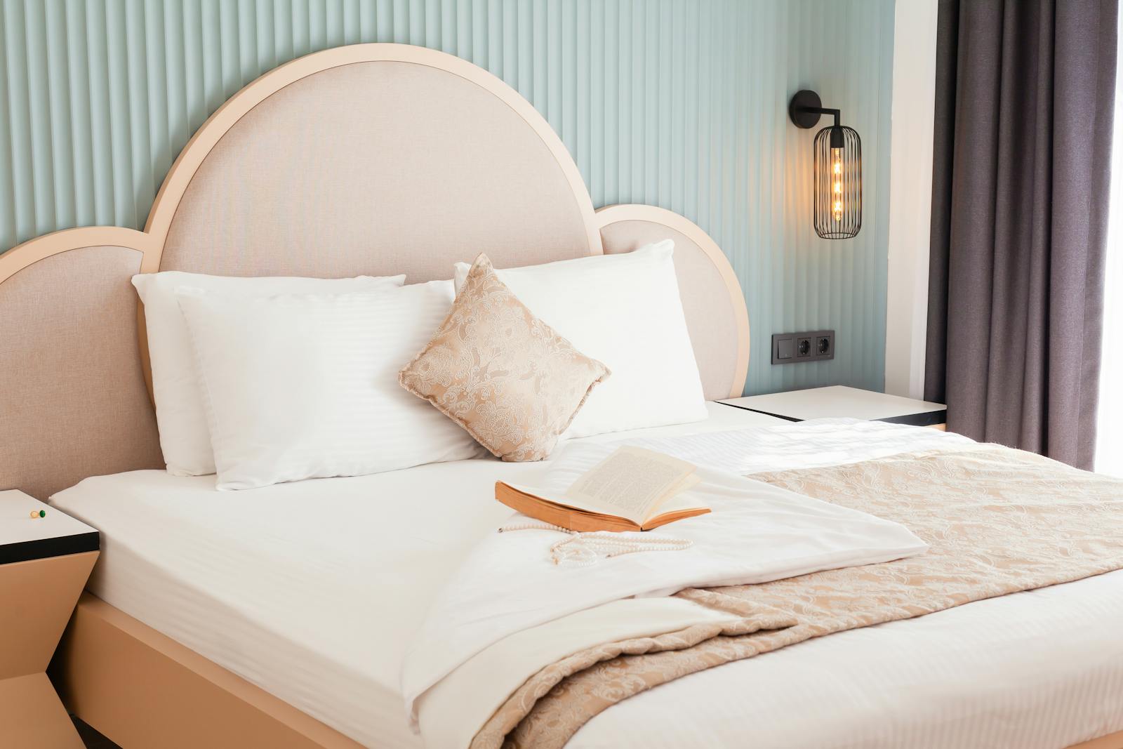 A made hotel bed with carefully arranged pillows and an open book - the calm of a room waiting for an expected guest.